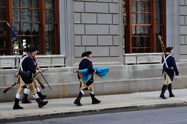 alt="שחקנים=מחופשים-לחיילי-המהפכה-האמריקאית-צועדים-ברחוב-בבוסטון">