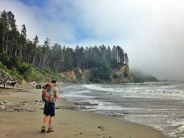alt="בחור-צועד-על-החוף-עם-תרמיל-וברקע-סלעים-מיוערים-הפארק-הלאומי-אולימפיק">