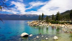 צילום נוף של אגם טאהו (Lake Tahoe) עם מים בצבע טורקיז וסלעי גרניט גדולים בקו החוף על רקע הרים מושלגים.