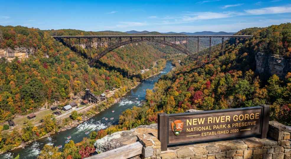 שלט עץ של הפארק הלאומי ערוץ הנהר החדש (New River Gorge National Park & Preserve) בחזית נקודת תצפית, ברקע גשר ערוץ הנהר החדש (New River Gorge Bridge) הענק החוצה את הקניון המיוער בצבעי שלכת.