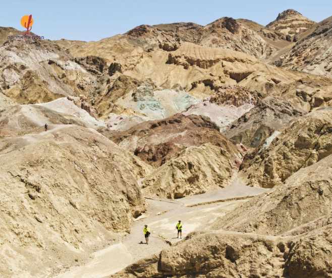 שני מטיילים בחולצות זוהרות צועדים בלב קניון הזהב (Golden Canyon) בעמק המוות, מוקפים בנקיקים וגבעות סלע מוזהבות וצבעוניות.