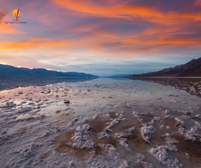 תצלום פנורמי מרהיב של שקיעה מעל אגן באדווטר (Badwater Basin) בעמק המוות, המציג את משטחי המלח המשושים המשתקפים במים הרדודים.