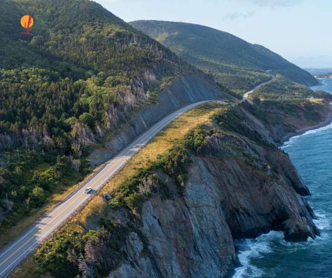 צילום אוויר פנורמי של שביל קאבוט (Cabot Trail) בנובה סקוטיה, קנדה. הכביש המפותל חצוב בצוקים דרמטיים מעל האוקיינוס האטלנטי, כשמימין נראים גלים כחולים מתנפצים. משמאל, הרים תלולים מכוסים ביערות ירוקים צפופים. מכונית בודדה נוסעת על הכביש, המציע נוף עוצר נשימה. בפינה השמאלית העליונה מופיע לוגו קטן של "מגלים את אמריקה".